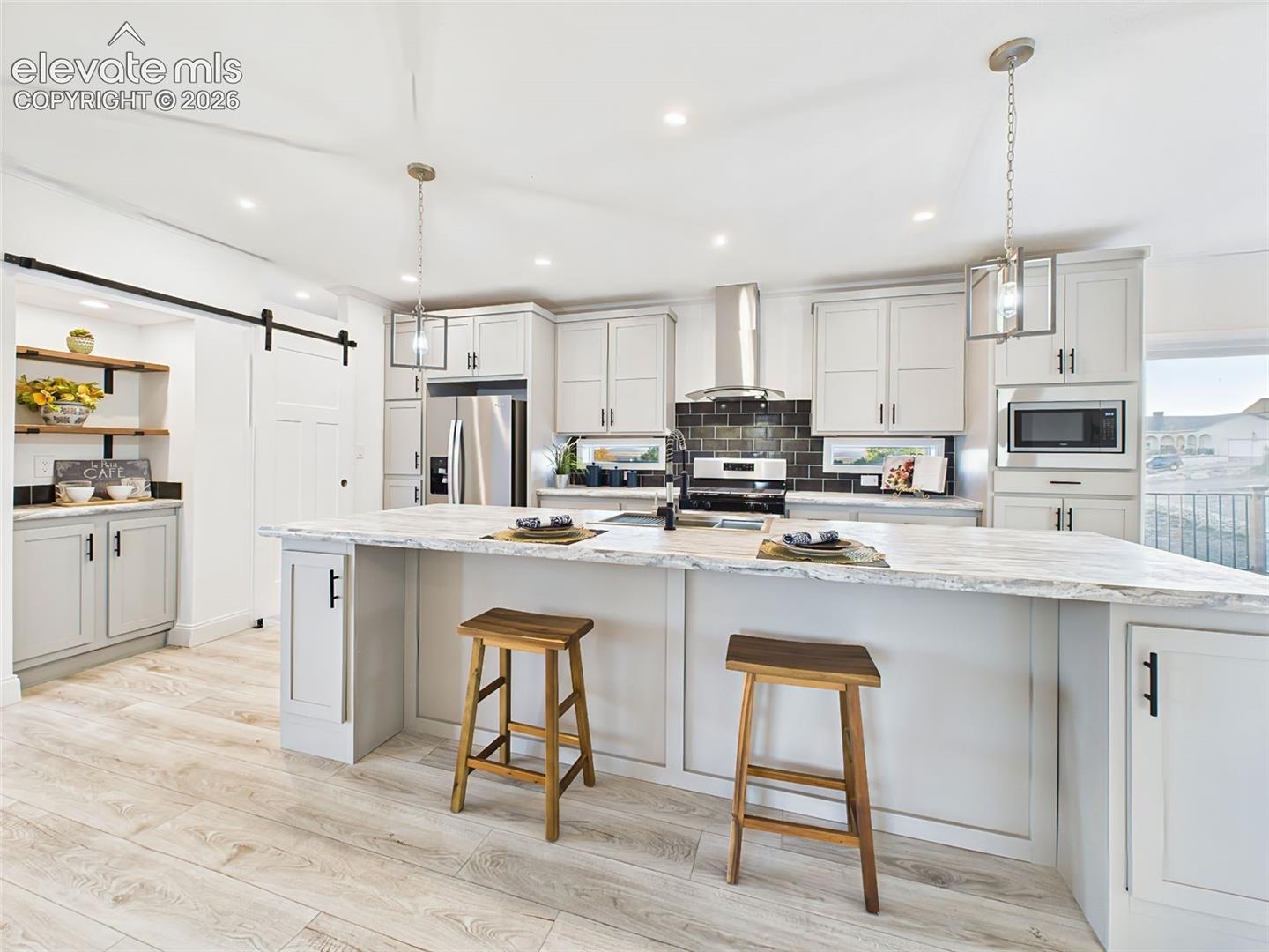 Spacious kitchen with an oversized center island, abundant cabinetry, stainless steel appliances, pendant lighting, and a distinctive dark tile backsplash. A sliding barn door provides access to the walk-in pantry.