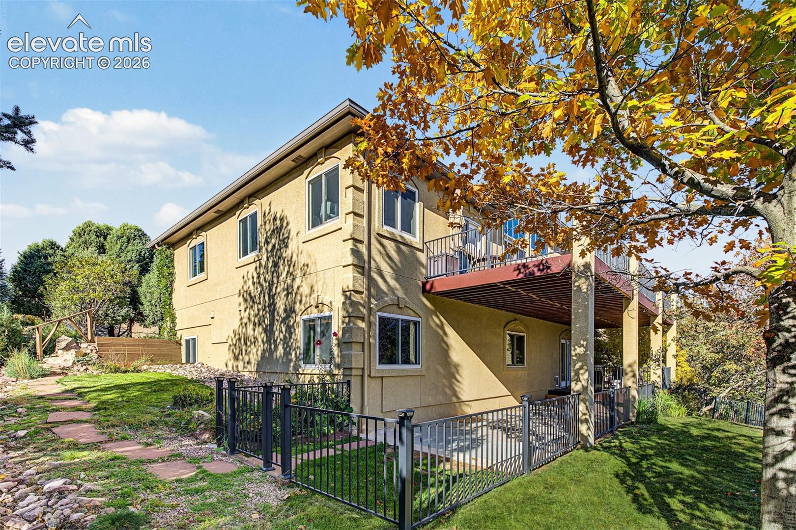 Rear exterior view showcasing the main-level balcony, covered patio below with direct access to the walkout basement, fenced yard area, and landscaped grounds with mature trees, natural stone accents, and a pathway along the side of the home.