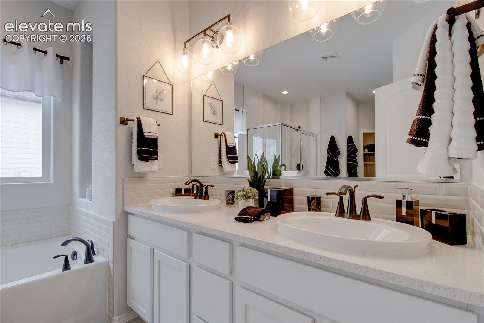 Dual vanity featuring white shaker cabinetry, quartz countertops, undermount sinks, and oil-rubbed bronze fixtures, complemented by a subway tile backsplash and a deep soaking tub