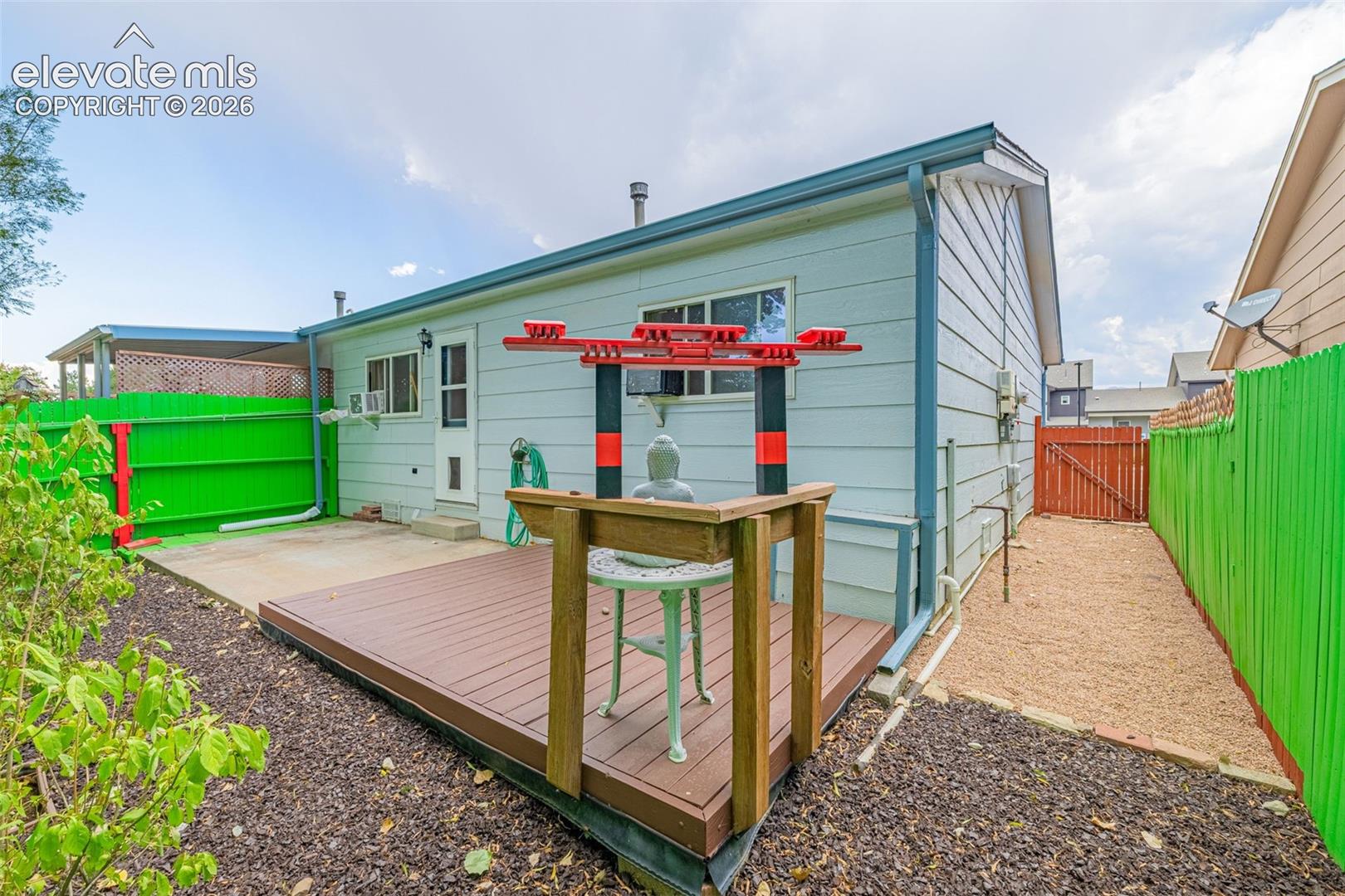 Rear exterior featuring light blue siding, a wood deck, concrete patio, and a gravel pathway