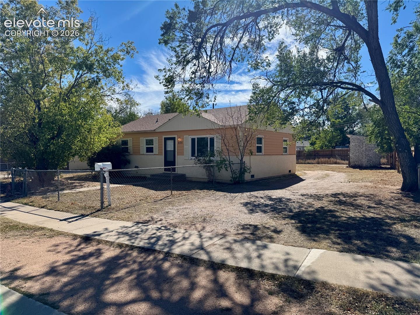Single-story exterior featuring dual-tone siding, a gable roof, chain-link perimeter fencing, and a detached storage structure