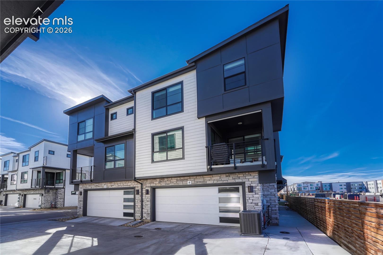 Contemporary multi-story exterior featuring a mix of horizontal siding, dark metal panels, and stone veneer accents, complete with private balconies, large windows, and an attached two-car garage with modern glass-paneled doors
