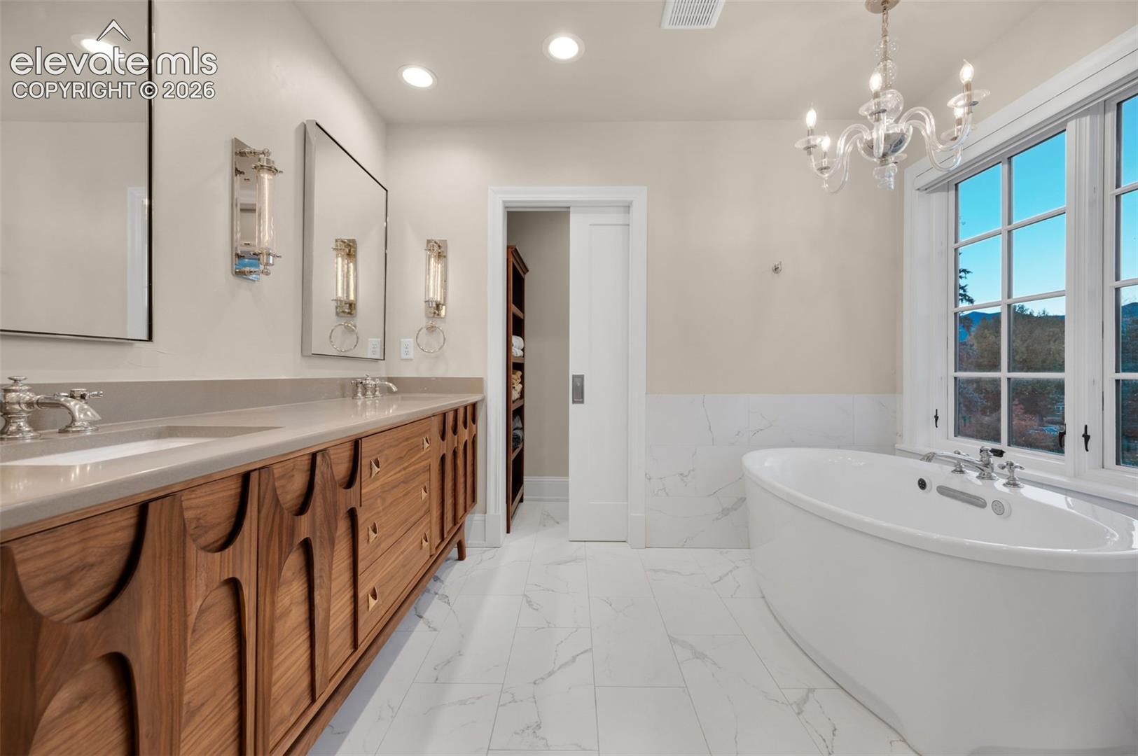 Bathroom featuring a wood-finish double vanity with stone countertops, a freestanding soaking tub, marble-patterned tile flooring, a tiered crystal chandelier, and a sliding door