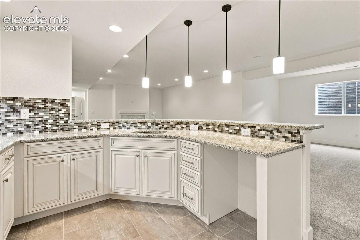 Kitchenette featuring off-white cabinetry, stone-finish countertops, decorative multi-tonal tile backsplash, recessed lighting, and three cylindrical pendant lights