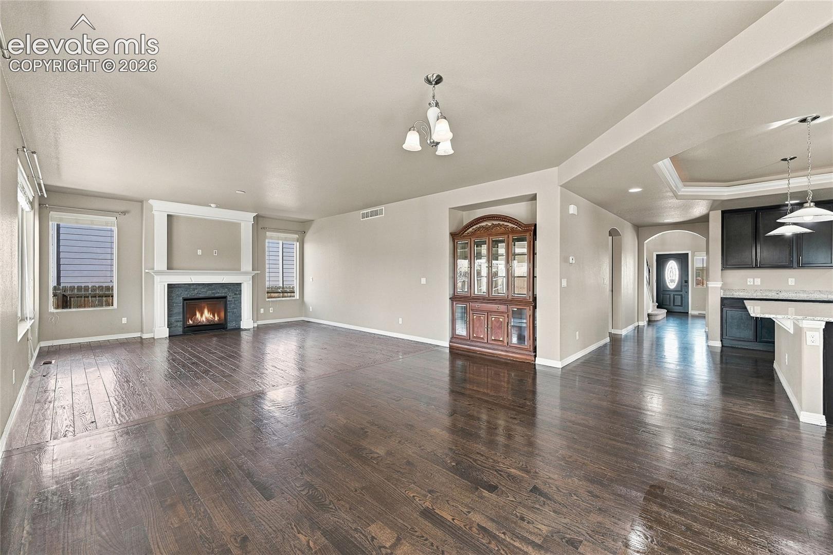 Open-concept area featuring wood-finish flooring, a fireplace with a stone surround, tray ceilings, recessed lighting, and built-in cabinetry