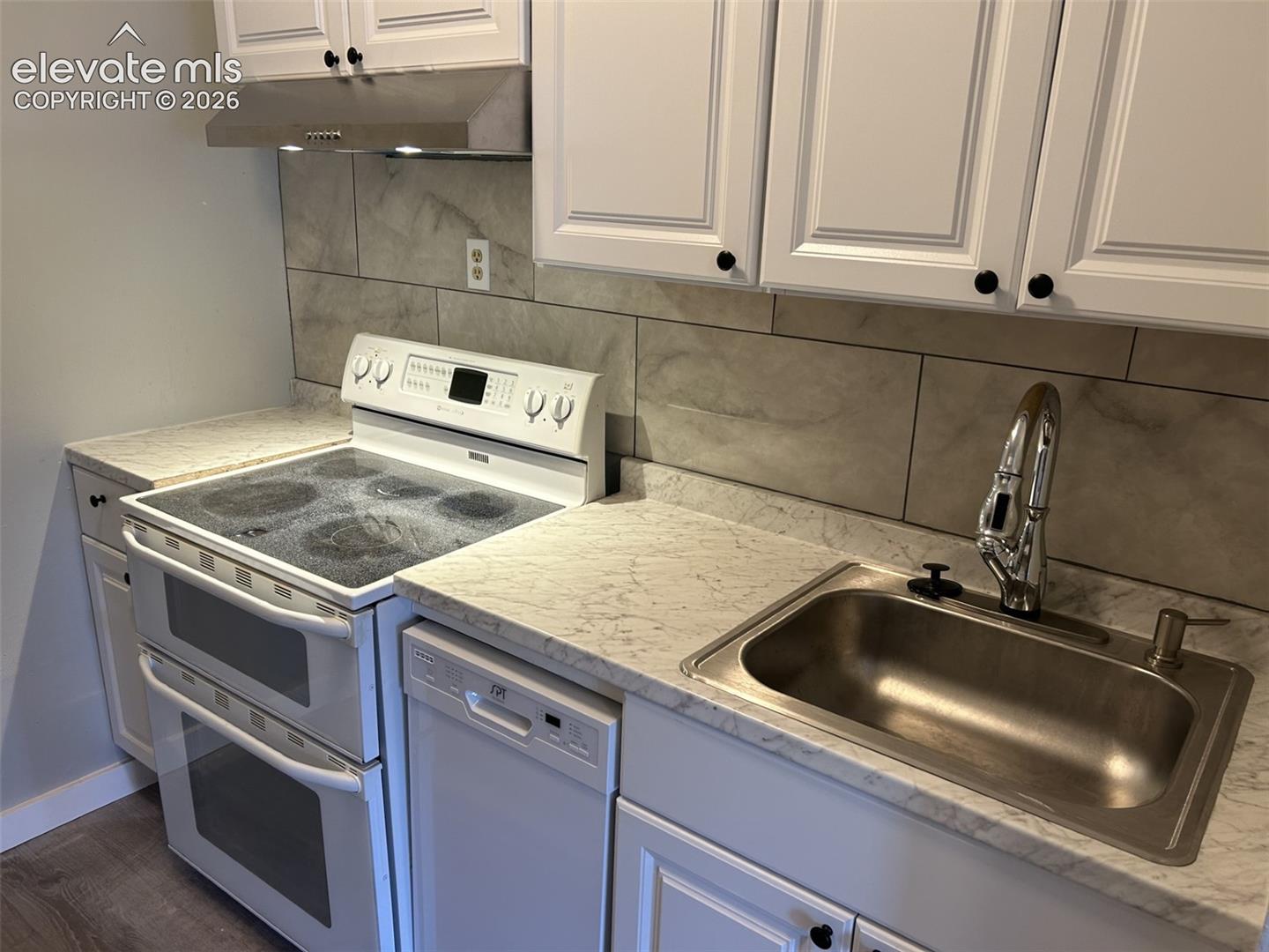 White shaker-style cabinetry with black hardware, stainless steel sink with a high-arc faucet, light-toned countertops, tile backsplash, and a double oven range