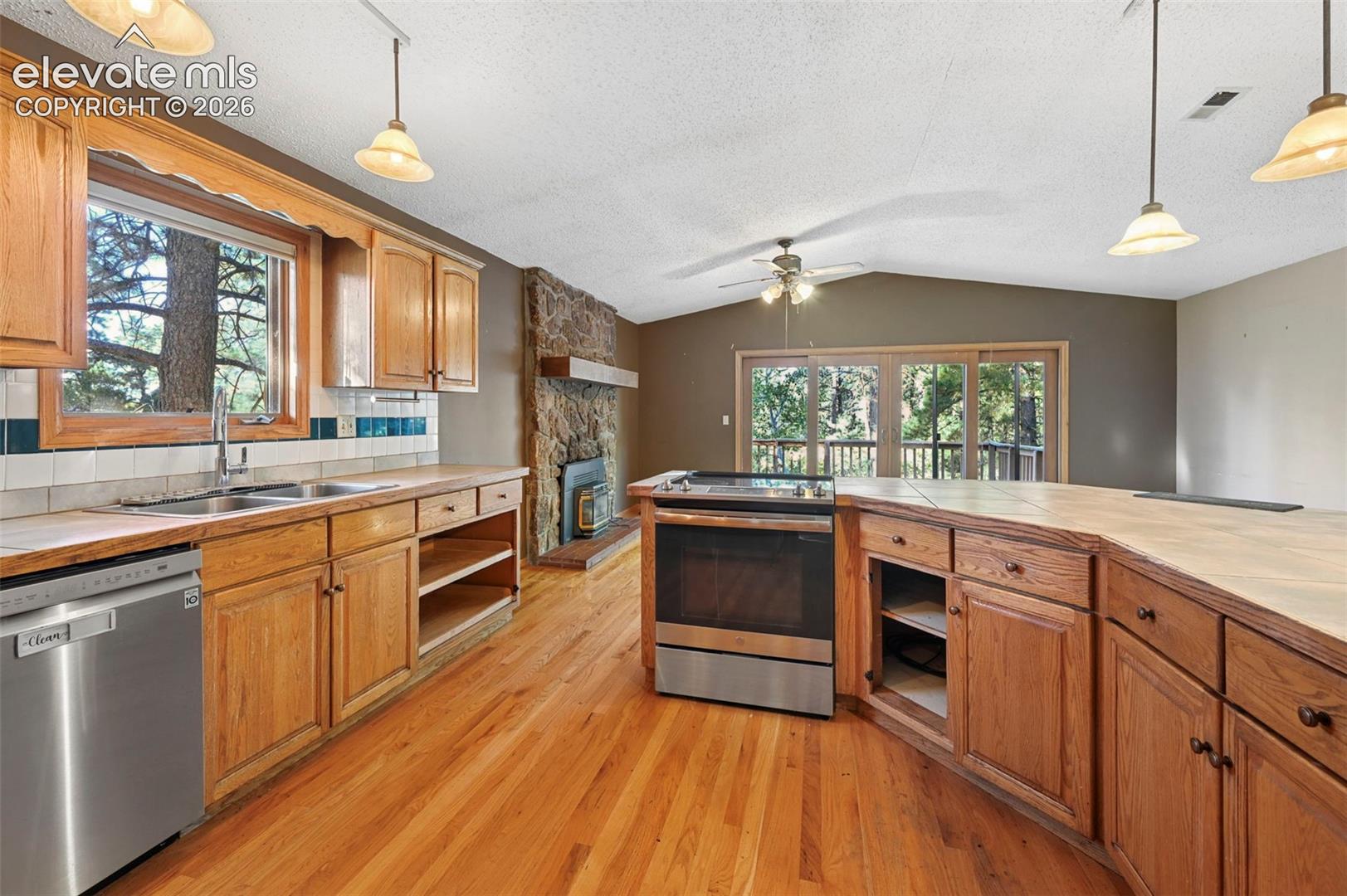 Hardwood flooring, wood-finish cabinetry, stainless steel dishwasher, stone-clad fireplace with mantel, and sliding glass doors with balcony access