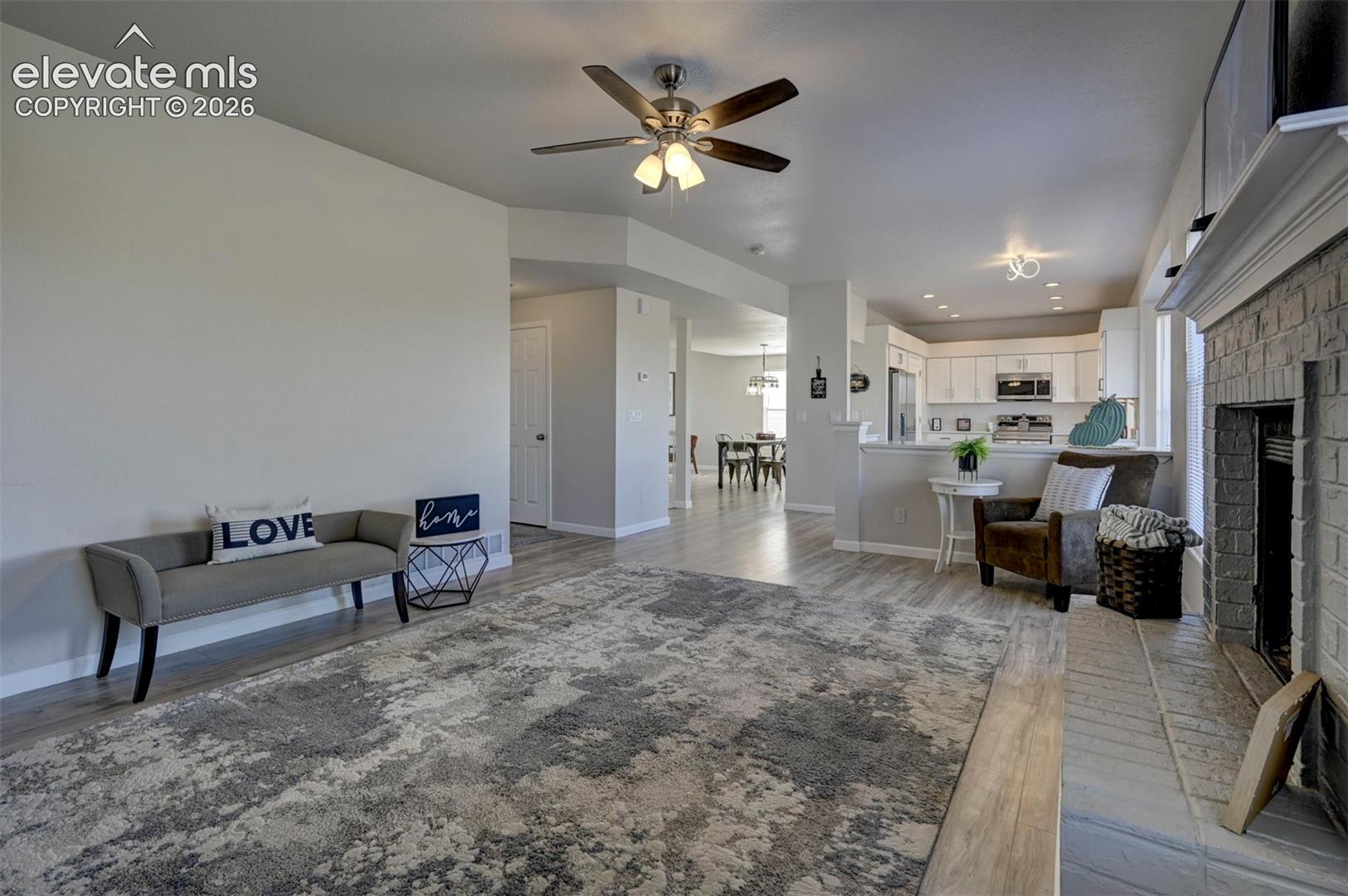 Living area featuring light-toned wood-finish flooring, a painted brick fireplace with a raised hearth, and a ceiling fan with integrated lighting