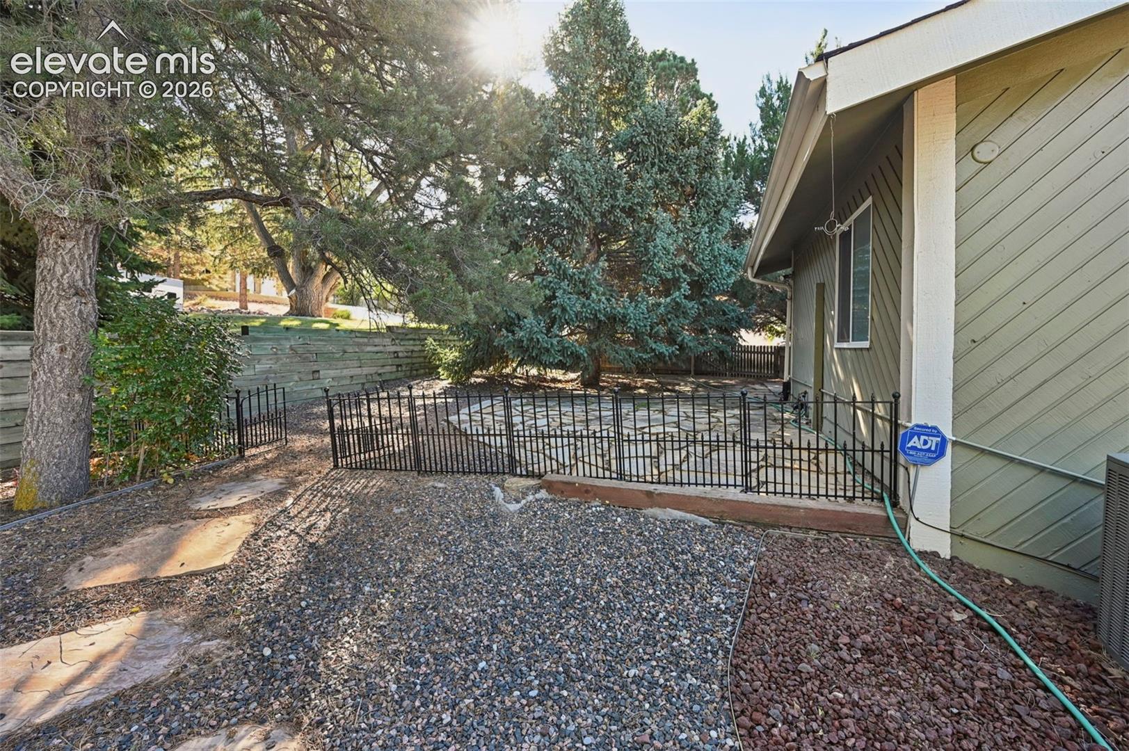 Backyard featuring a stone-paver patio, black metal fencing, timber retaining walls, mature pine trees, and gravel ground cover