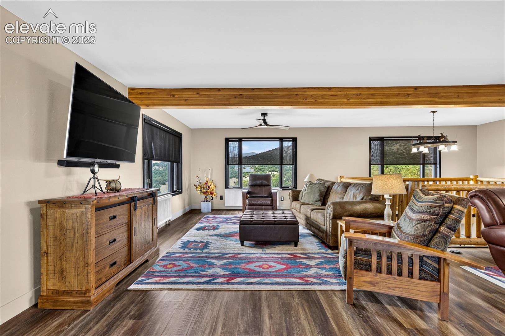Spacious living area featuring wood-finish flooring, exposed ceiling beams, and multiple windows