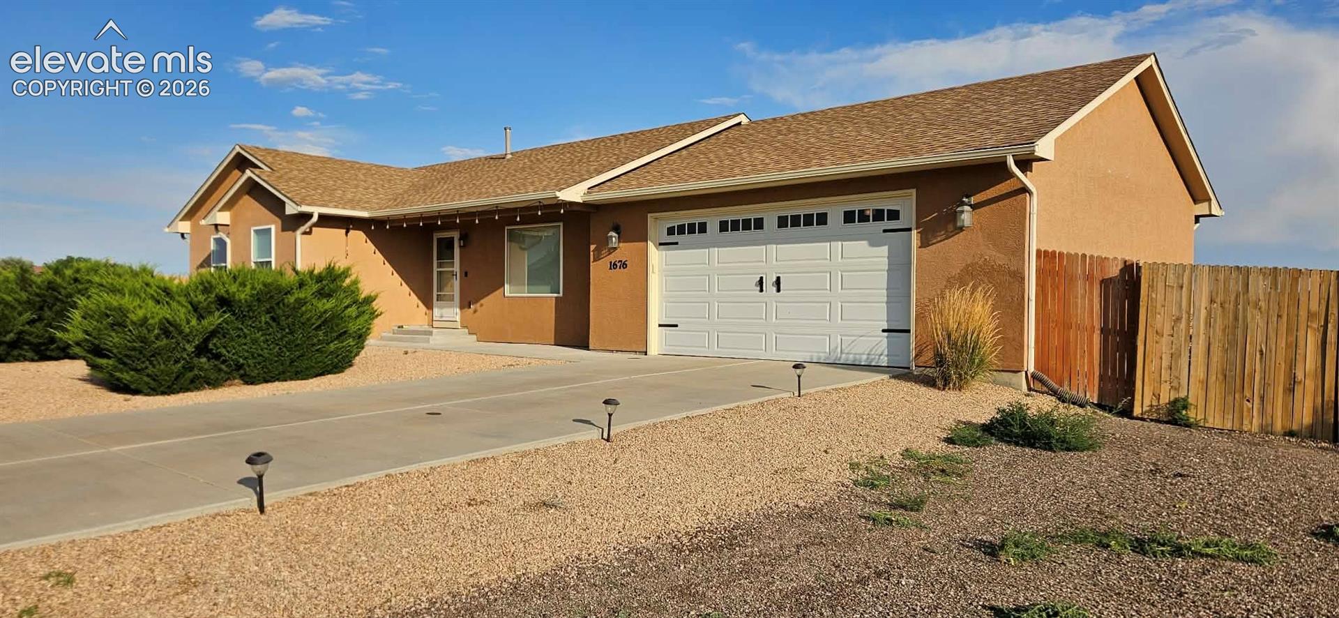 Single-story stucco exterior featuring a gabled roof, attached two-car garage with decorative hardware, concrete driveway, and rock-based landscaping