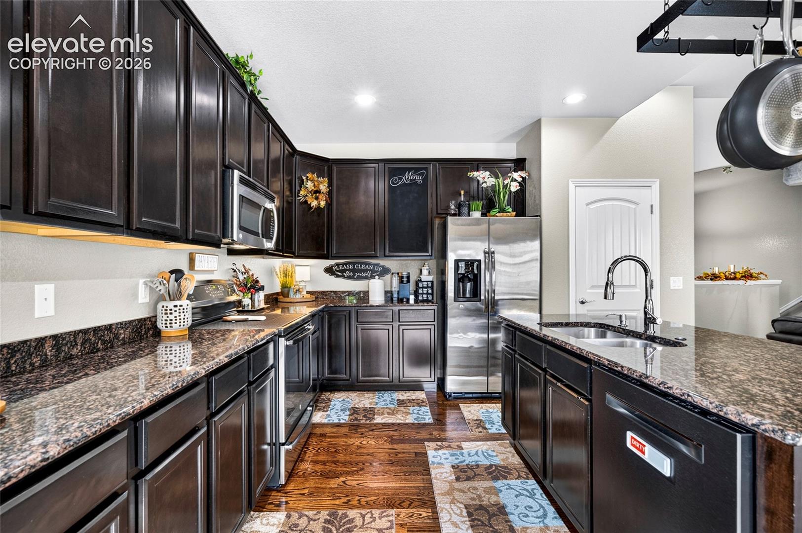 Kitchen featuring espresso-toned cabinetry, stone countertops, stainless steel appliances, a central island with a dual-basin sink, and wood-finish flooring