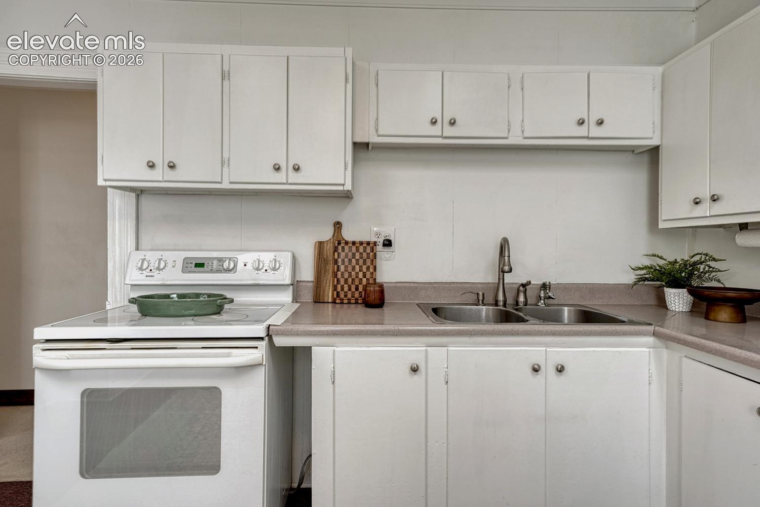White cabinetry with silver hardware, dual basin sink with gooseneck faucet, white electric range, and light-toned countertops