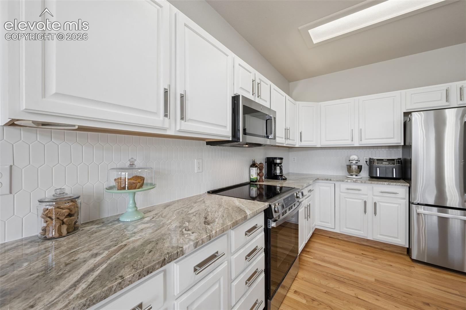 Kitchen featuring white cabinetry, stone-patterned countertops, a white hexagonal tile backsplash, stainless steel appliances, and wood-finish flooring