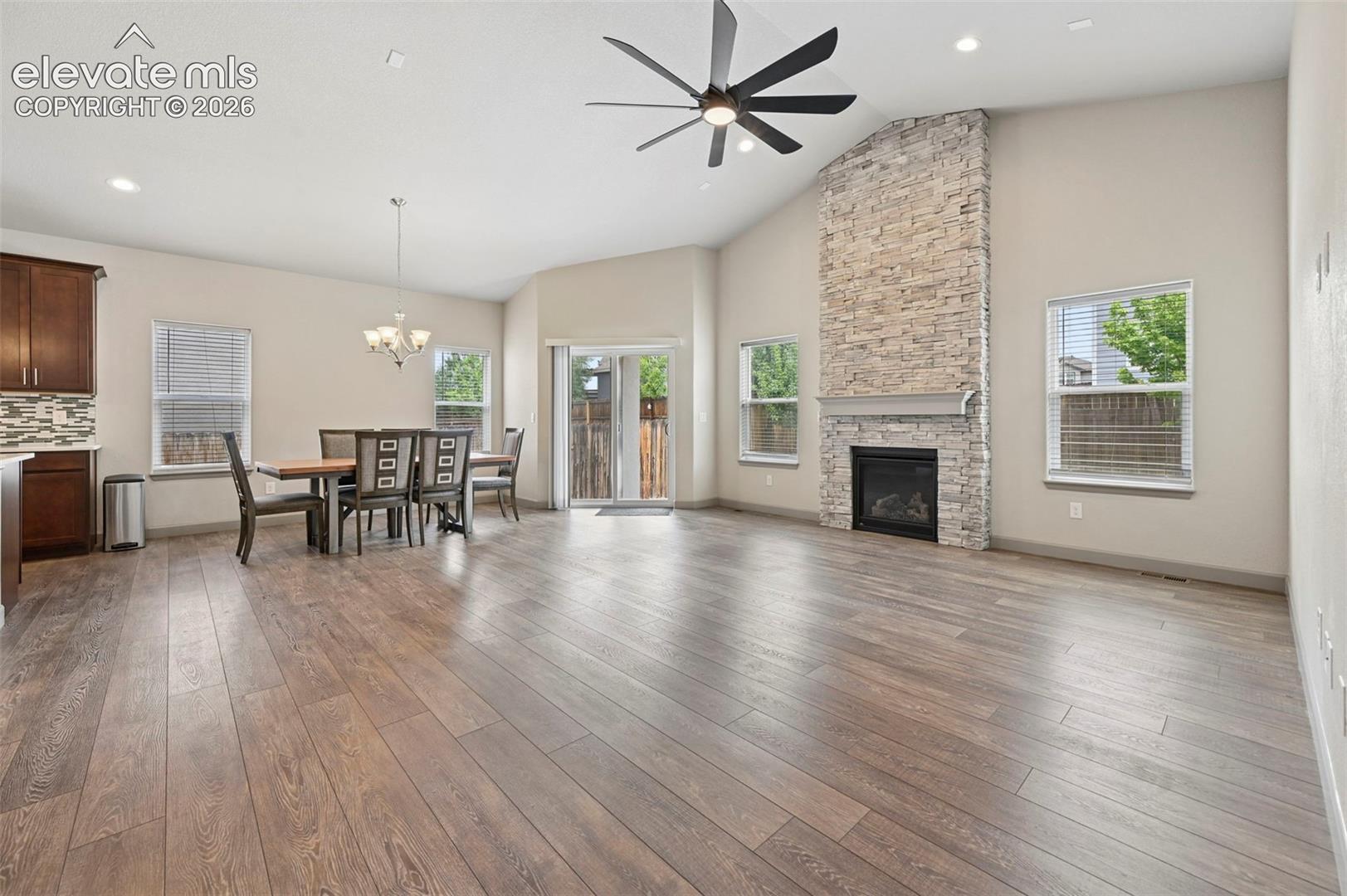 Open-concept living space featuring wood-finish flooring, vaulted ceilings, stone-surround fireplace, multi-blade ceiling fan, and sliding glass doors