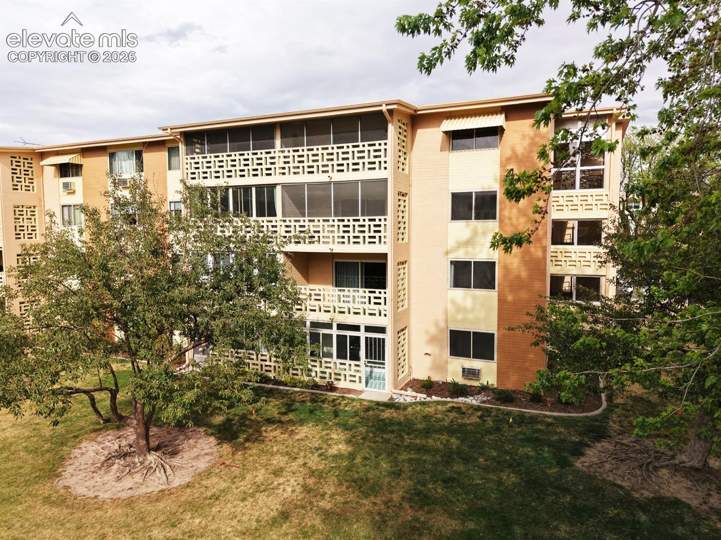 Multi-story building exterior featuring brick and stucco finishes, multiple balconies with decorative concrete screen walls, and a manicured lawn with mature trees