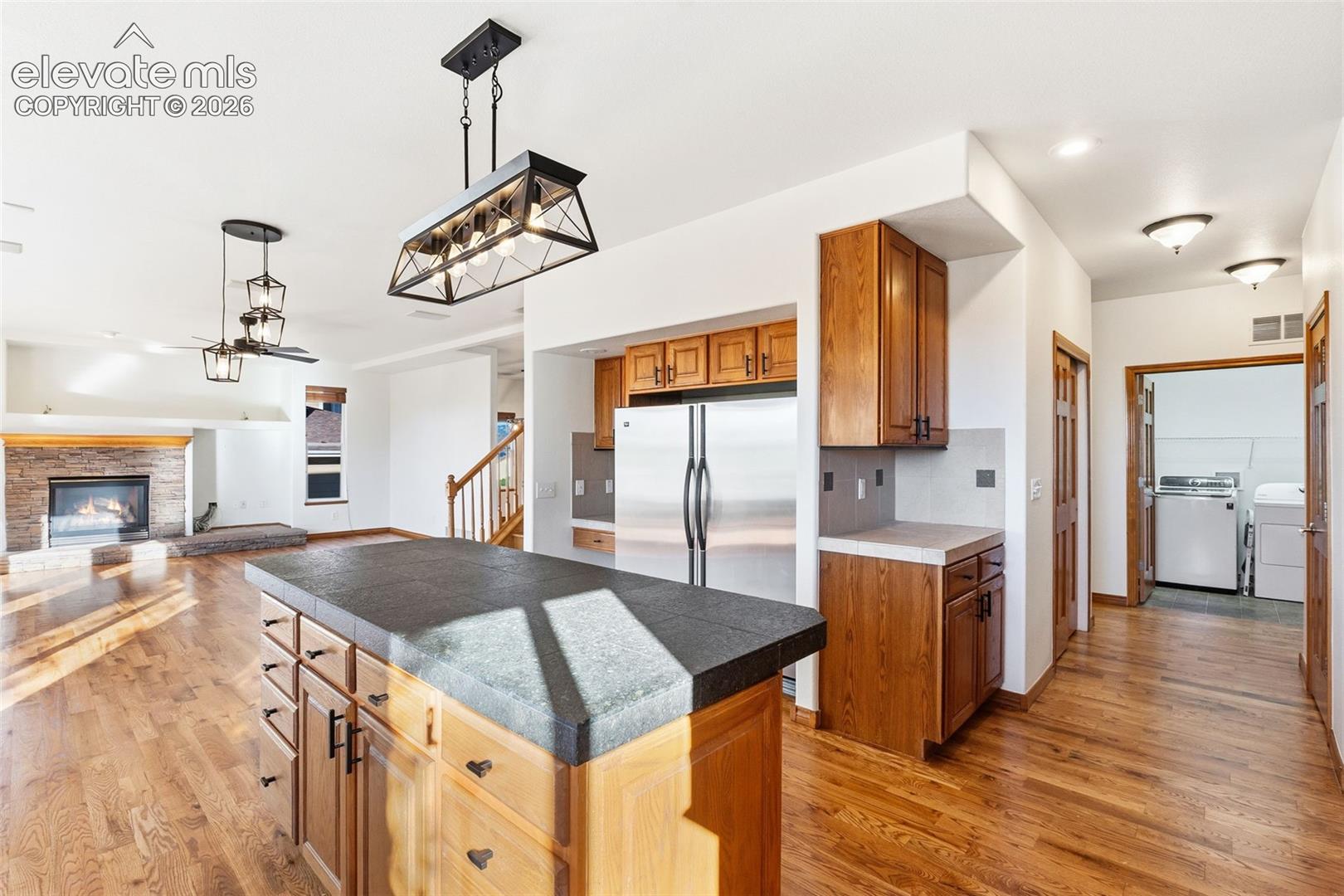 Kitchen featuring a central island with a stone-finish countertop, wood-finish cabinetry, stainless steel refrigerator, wood-finish flooring, and industrial-style pendant lighting