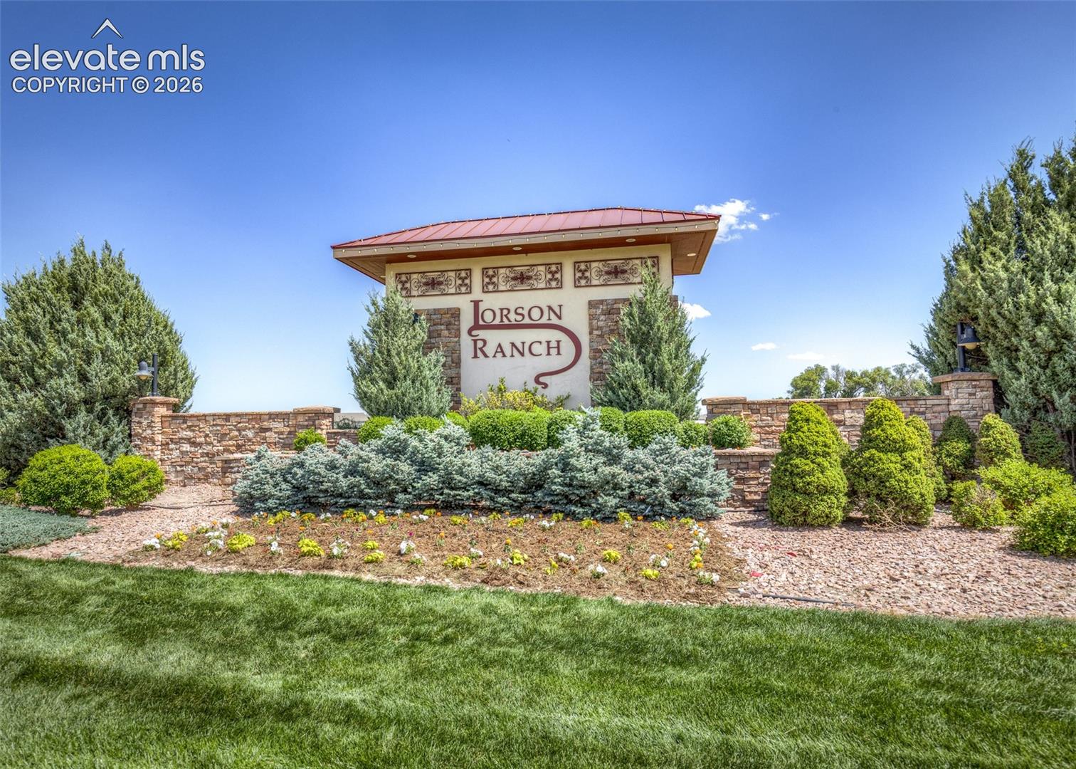 Community monument sign featuring stone masonry pillars, metal roof detailing, and professional landscaping with ornamental shrubs, gravel beds, and a grass lawn