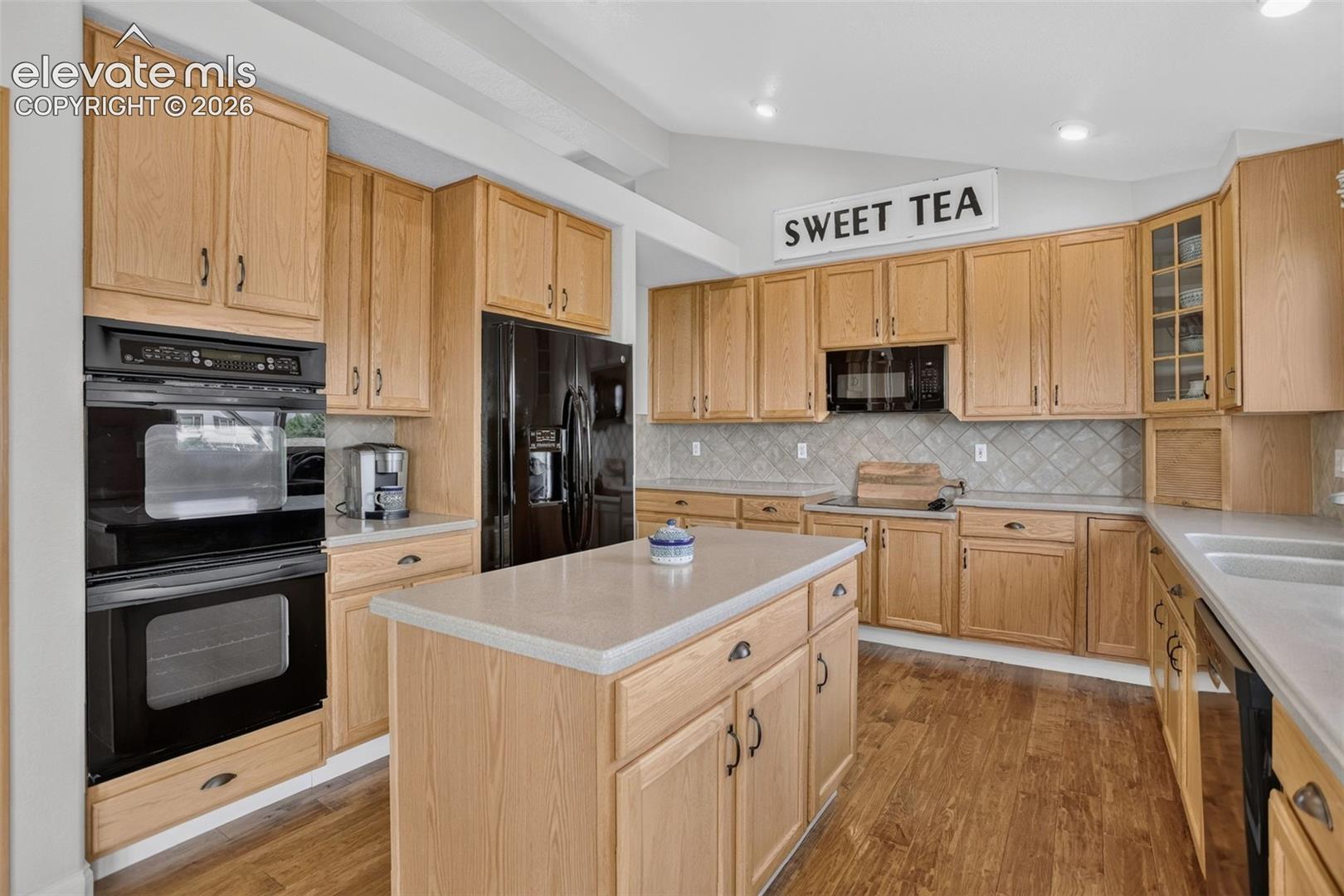 Spacious kitchen featuring light wood cabinetry, a center island with countertop, black built-in appliances, and wood-finish flooring