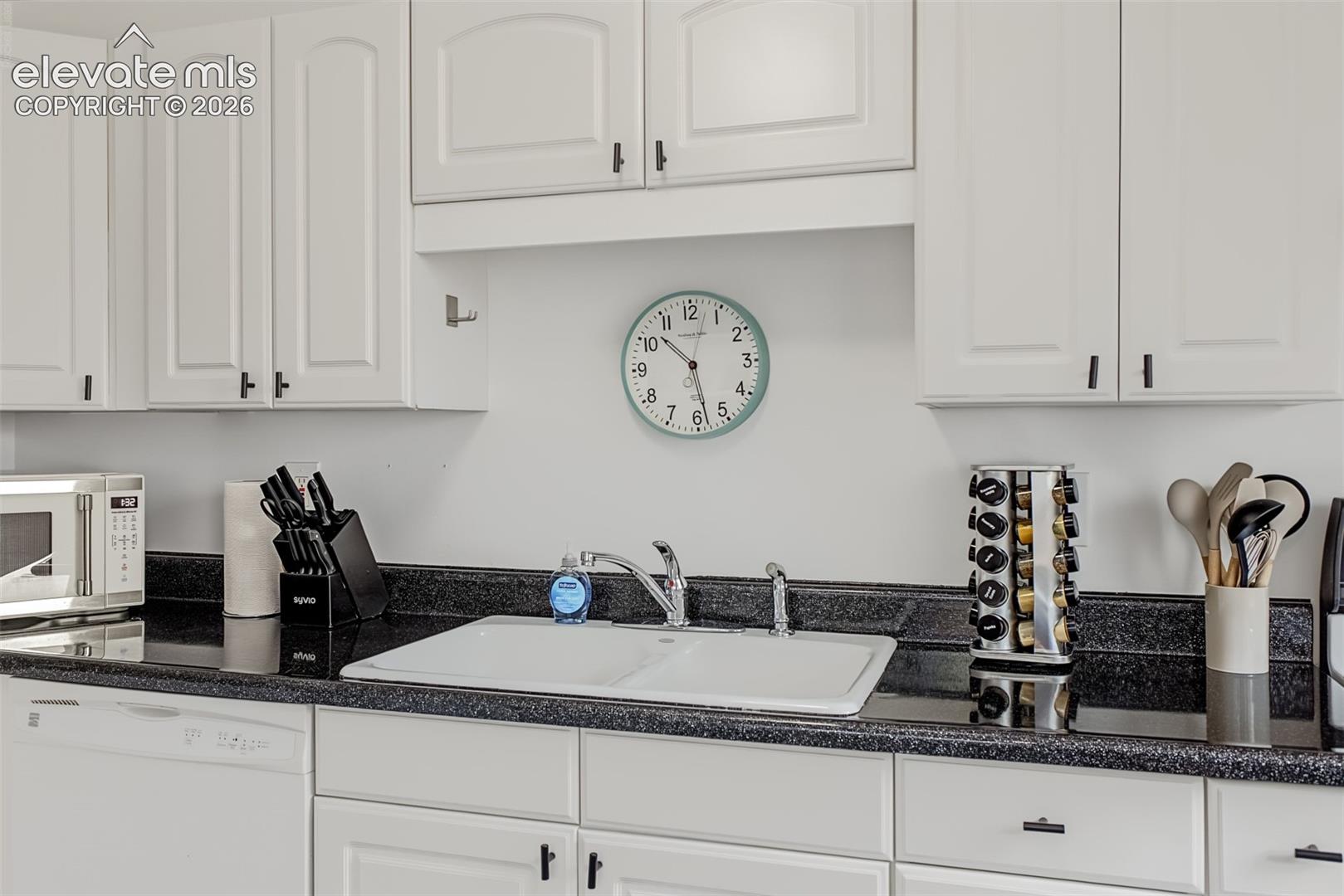 White shaker-style cabinetry, dark stone-finish countertops, dual-basin white sink, and chrome-finish faucet hardware