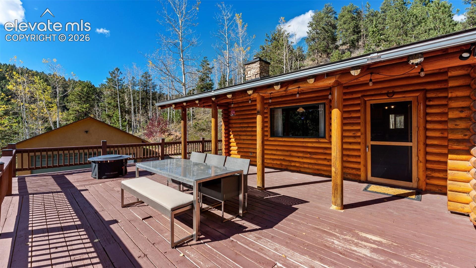 Covered log-walled deck featuring timber support columns, wood decking, metal railing, and stone chimney masonry