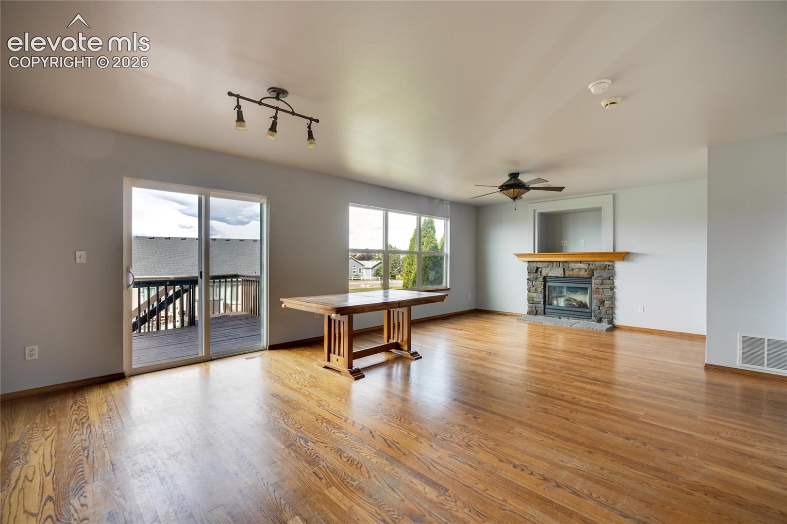 Hardwood flooring, sliding glass door to exterior deck, stone fireplace with wood mantel, multiple windows, and a ceiling fan