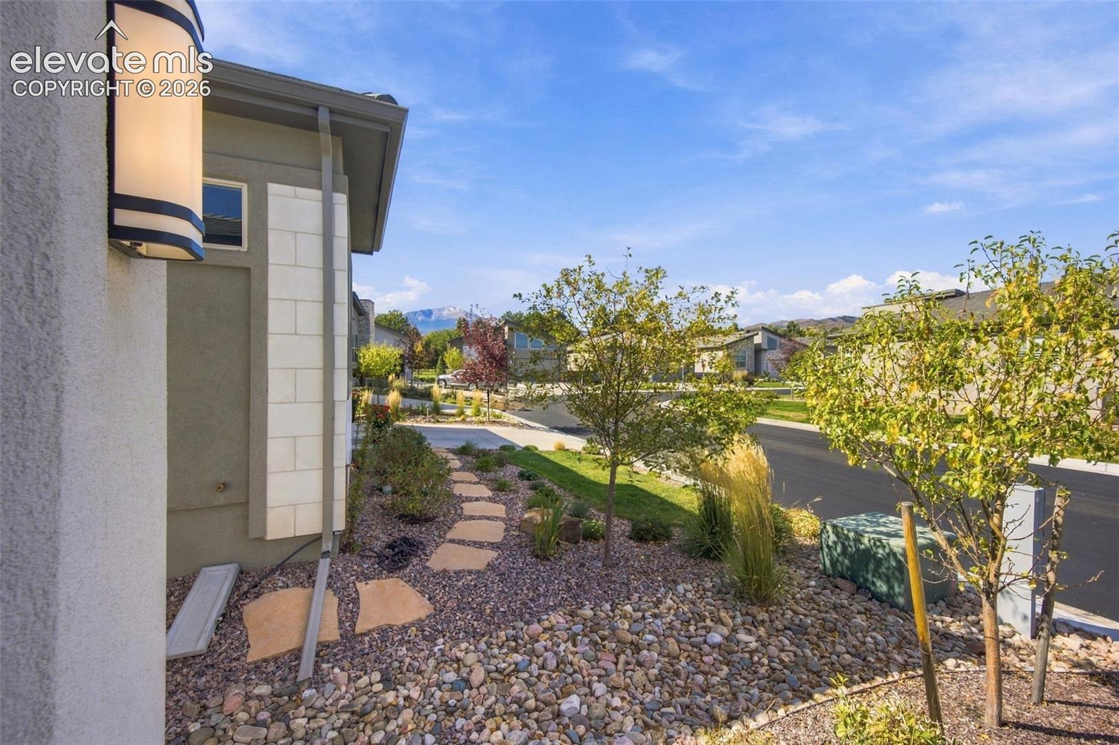 Exterior wall featuring light-toned stone accents, wall-mounted sconce lighting, stone paver pathway, decorative rock landscaping, and mature ornamental trees with mountain scenery