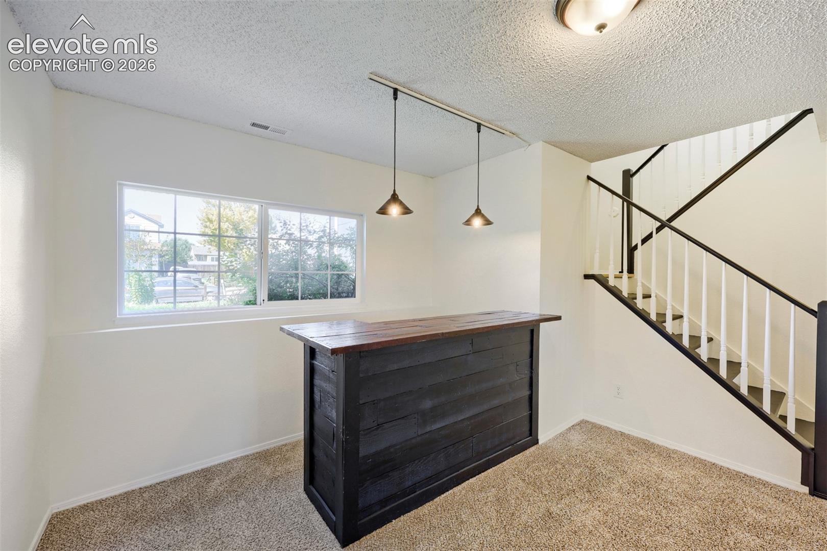 Built-in bar with wood-finish countertop, track lighting with pendant fixtures, carpeted flooring, and a staircase with white spindles and dark wood-finish handrails