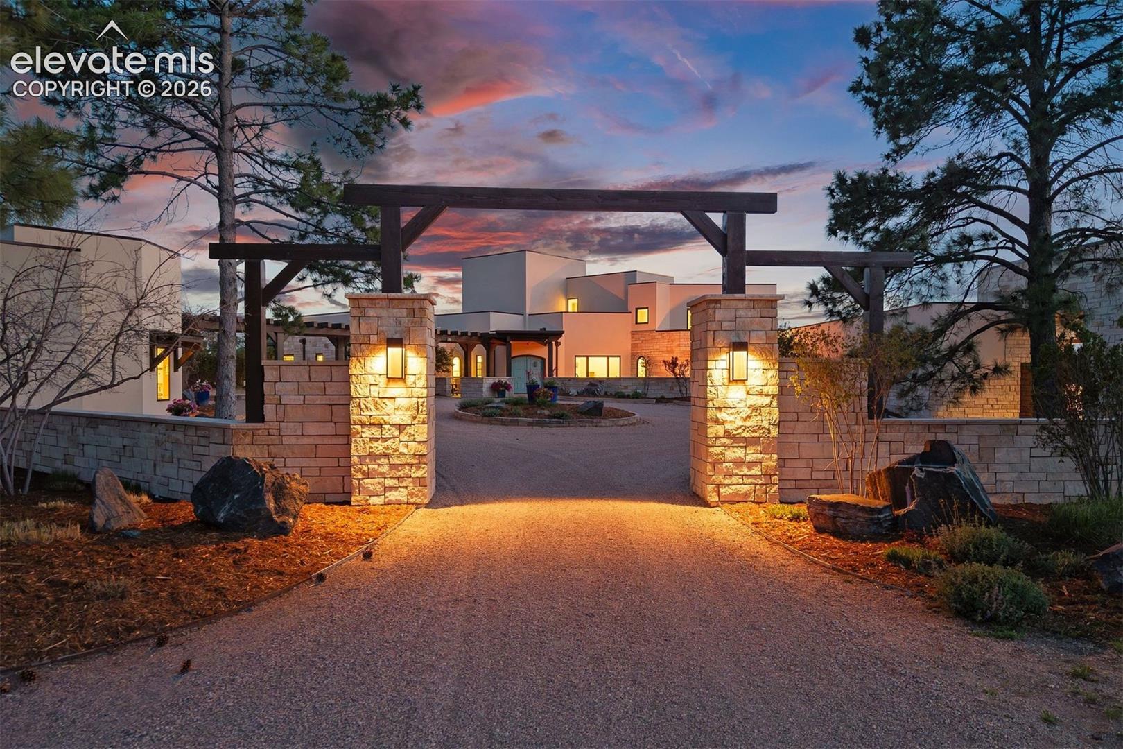 The driveway passes through a dramatic gated entry with stacked stone pillars and a timber beam overhead, opening into a circular motor court centered by a landscaped island.