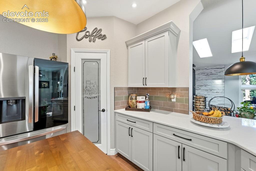 Stainless steel refrigerator, pantry door with frosted glass, light cabinetry with dark hardware, solid surface countertops, and a tiled backsplash
