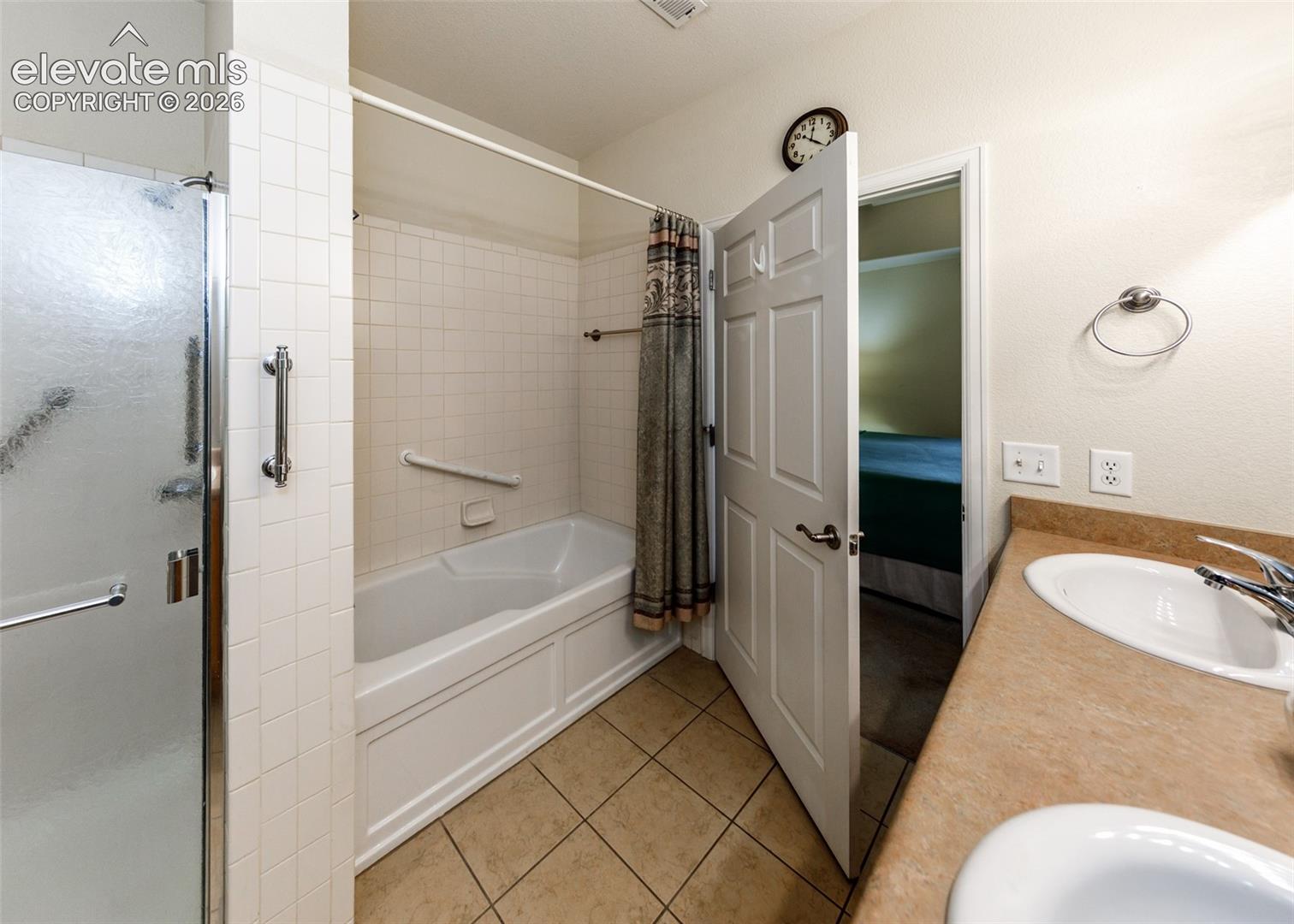 Bathroom featuring a dual-sink vanity with a solid-surface countertop, a separate shower enclosure with a glass door, a tile-surround soaking tub, and tile flooring