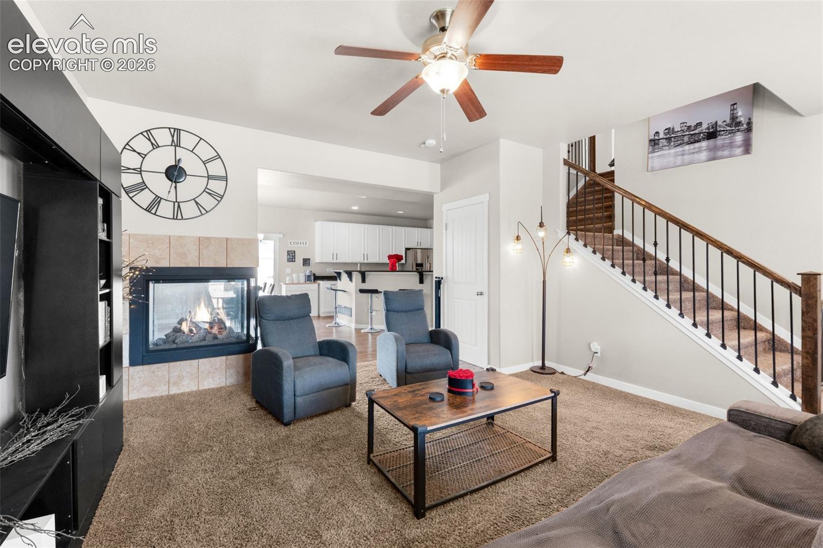 Living area featuring a double-sided gas fireplace with tile surround, built-in black shelving, a central ceiling fan, and a staircase with metal balusters