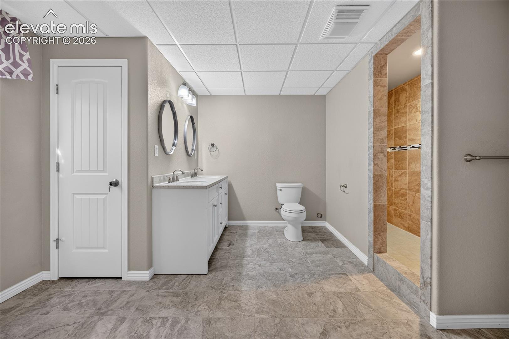 Dual-sink vanity with a neutral countertop, drop-in basin sinks, stone-tiled walk-in shower, and patterned floor tiling