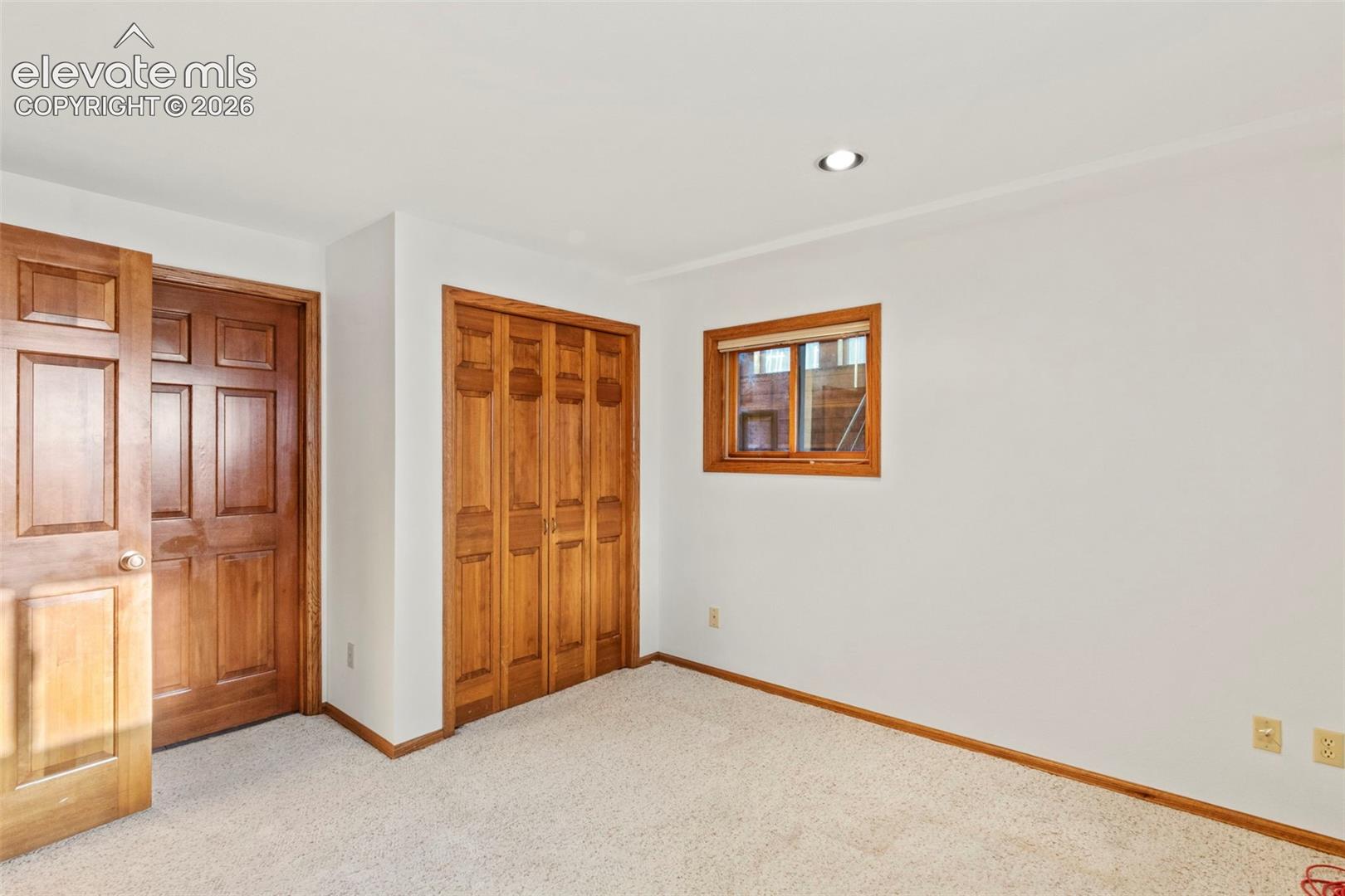 Room featuring neutral carpeting, six-panel wood-finish door, bi-fold wooden closet doors, recessed ceiling lighting, and a window with wood-finish trim