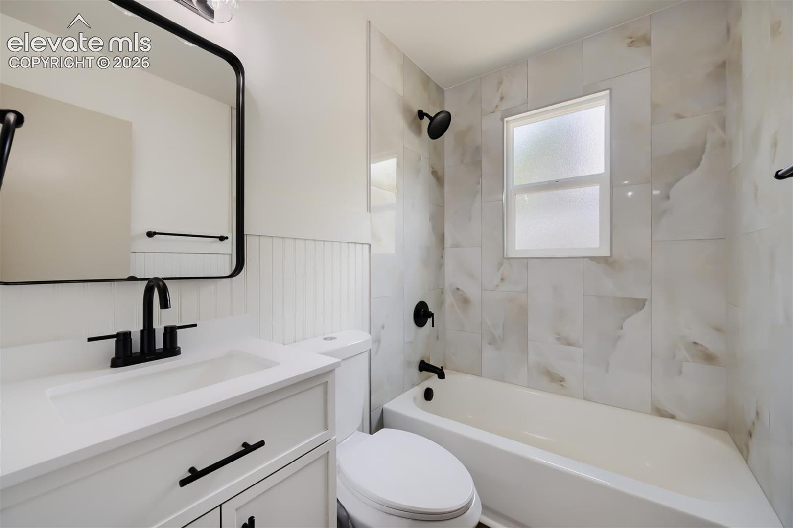 White vanity with solid surface countertop, matte black fixtures, full tile tub surround, and a wall-mounted mirror