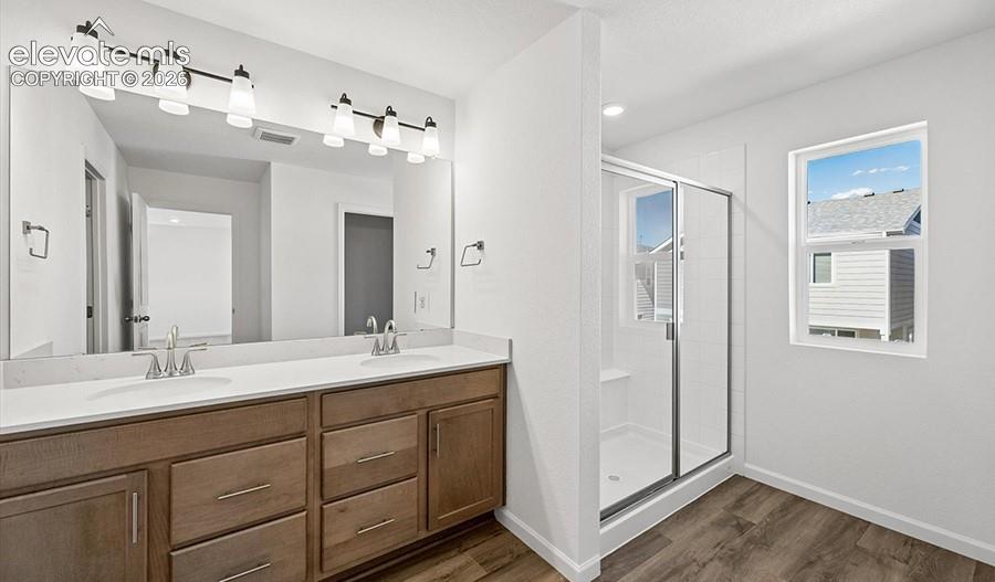 Dual-sink vanity with light-toned countertop, wood-finish cabinetry, full-width mirror, glass-enclosed shower, and wood-finish flooring