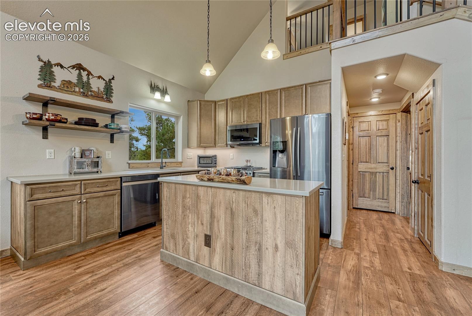 Kitchen featuring vaulted ceilings, wood-finish flooring, a central island with a solid surface countertop, stainless steel appliances, and wood cabinetry