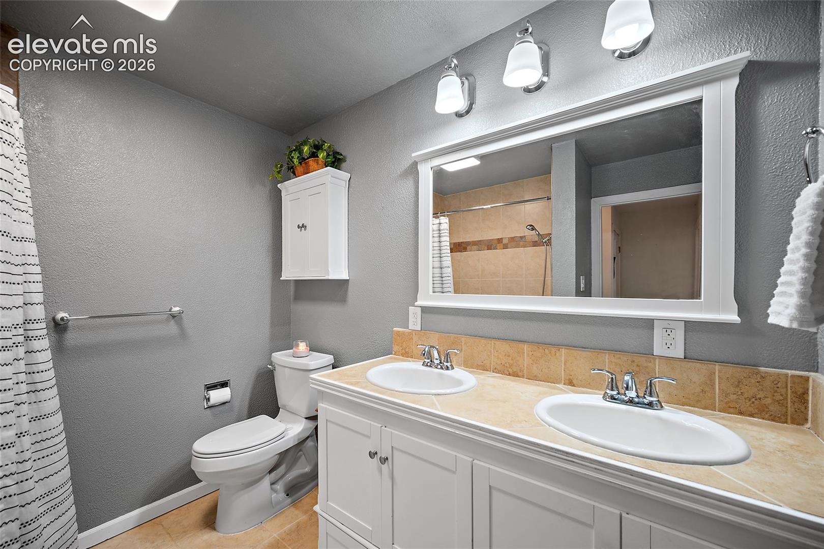 Double vanity featuring white cabinetry, a neutral stone-finish countertop, dual under-mount sinks, a wide-frame wall mirror, and three-light fixture