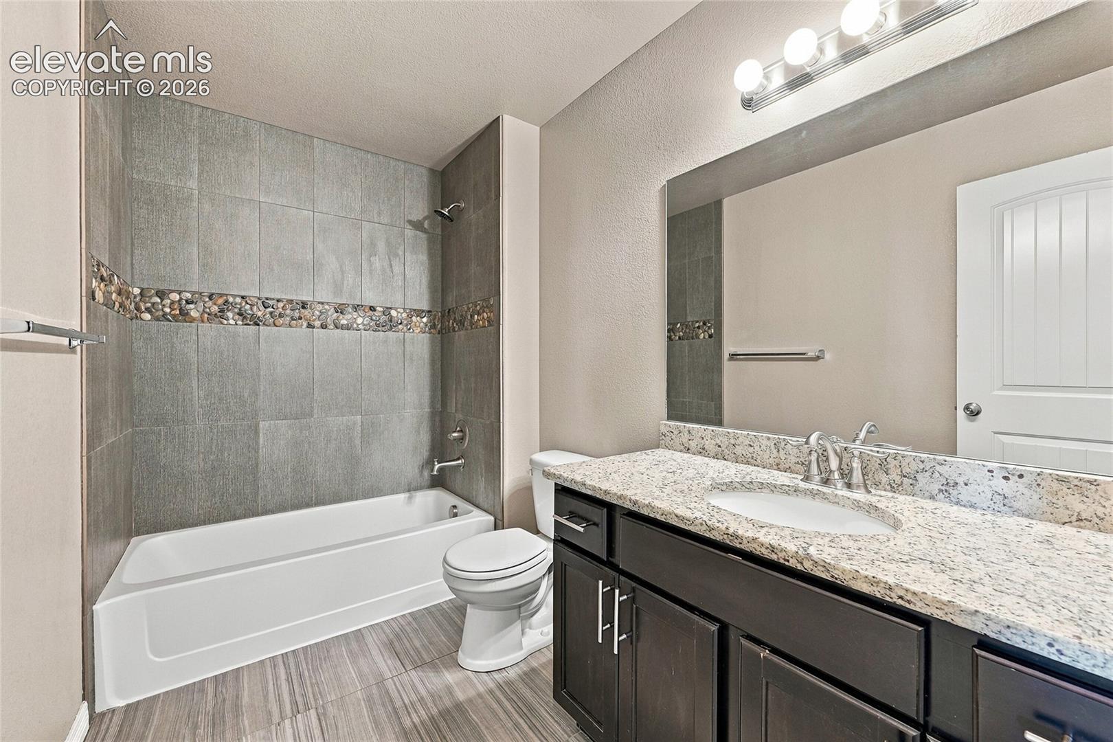 Bathroom featuring a vanity with stone-finish countertop, dark cabinetry, full-width wall mirror, bathtub with tiled surround, and decorative pebble-tile accent
