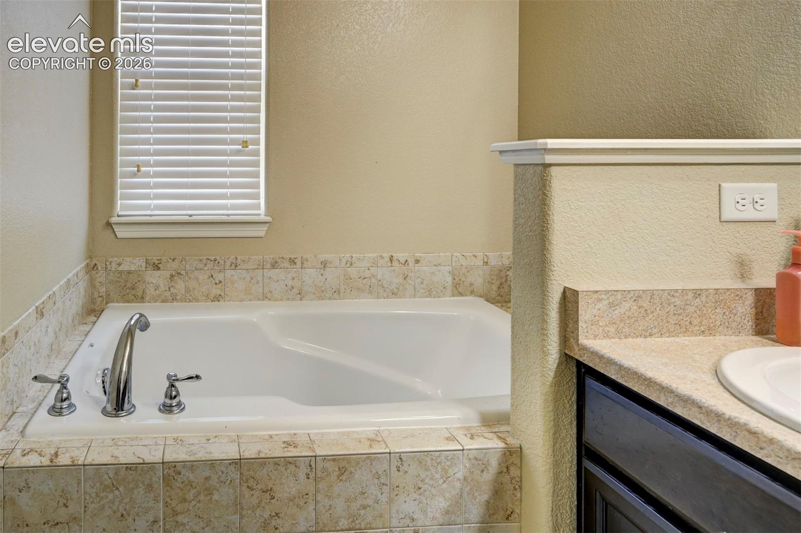 Built-in soaking tub with chrome fixtures, surrounded by neutral-toned tiled surfaces
