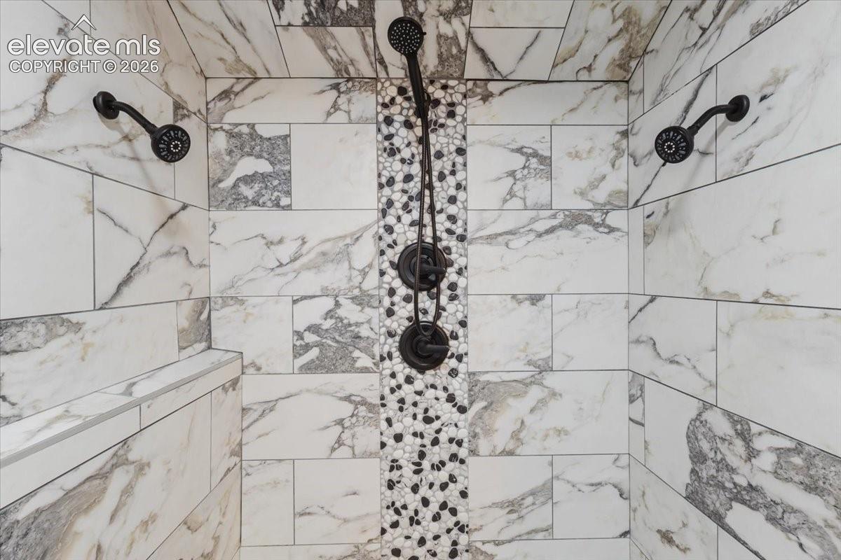 Shower enclosure featuring large format marble-look tile, pebble-style accent tile, three shower heads, and an integrated shower bench
