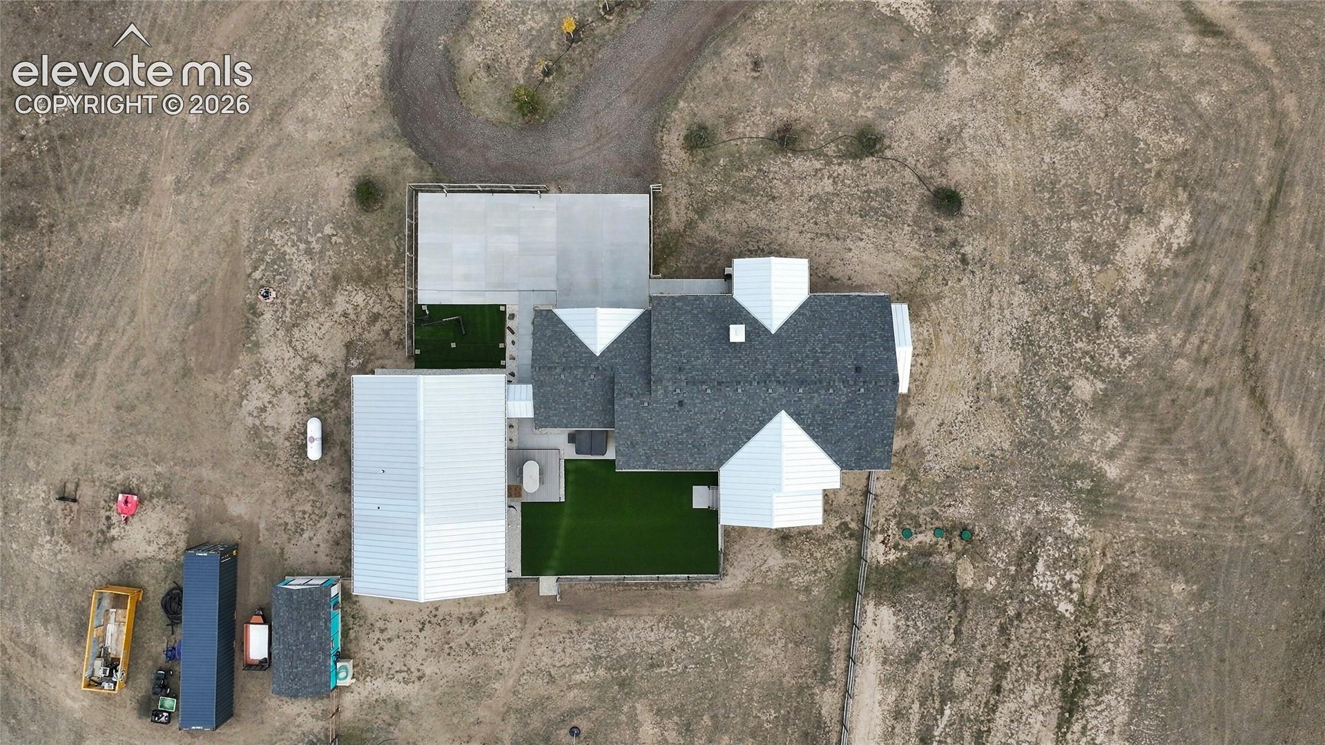 Aerial view of the property featuring a structured roofline with shingle and metal roofing, a winding gravel driveway, and a fenced artificial turf area