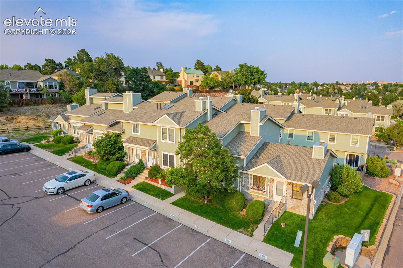 Residential complex featuring multiple units, ample surface parking, individual front entrances, manicured landscaping, and mature trees
