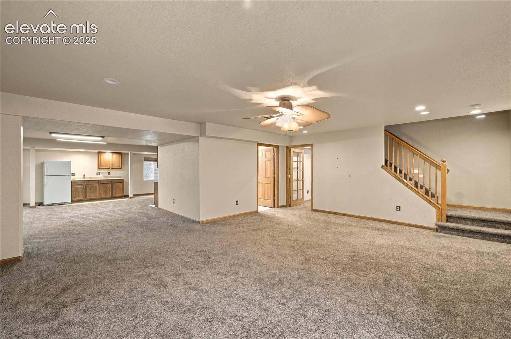 Open-concept interior featuring carpeting, a kitchenette with wood-finish cabinetry, a ceiling fan with integrated lighting, and a staircase with a wooden balustrade