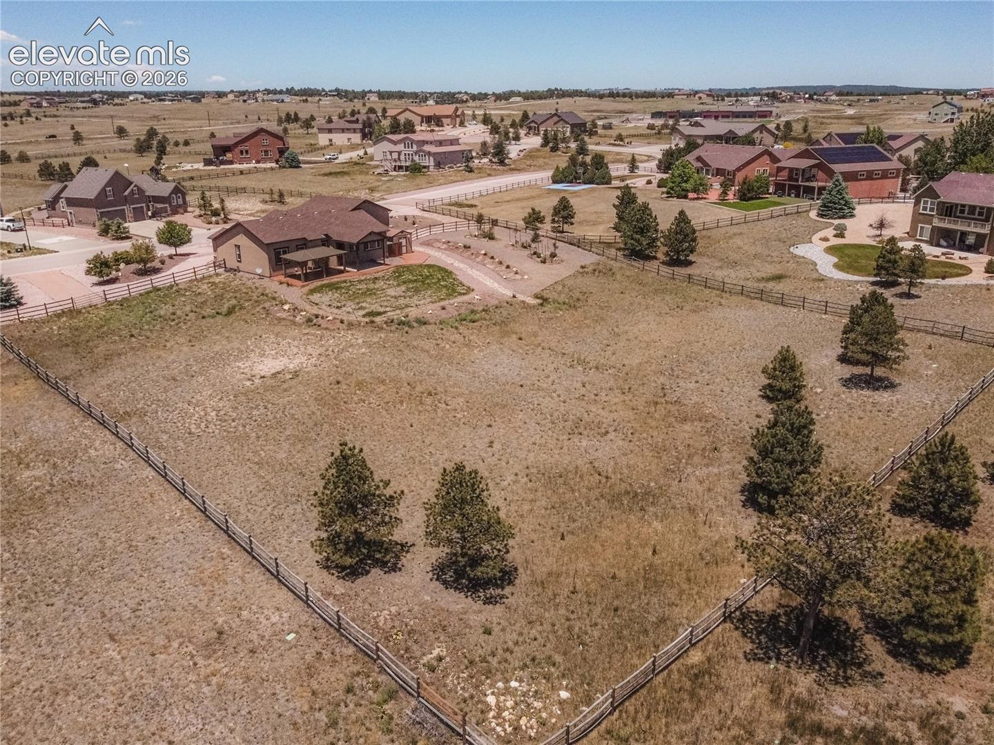 Expansive property featuring a large undeveloped lot, perimeter fencing, mature evergreen trees, and a residential structure with a covered patio