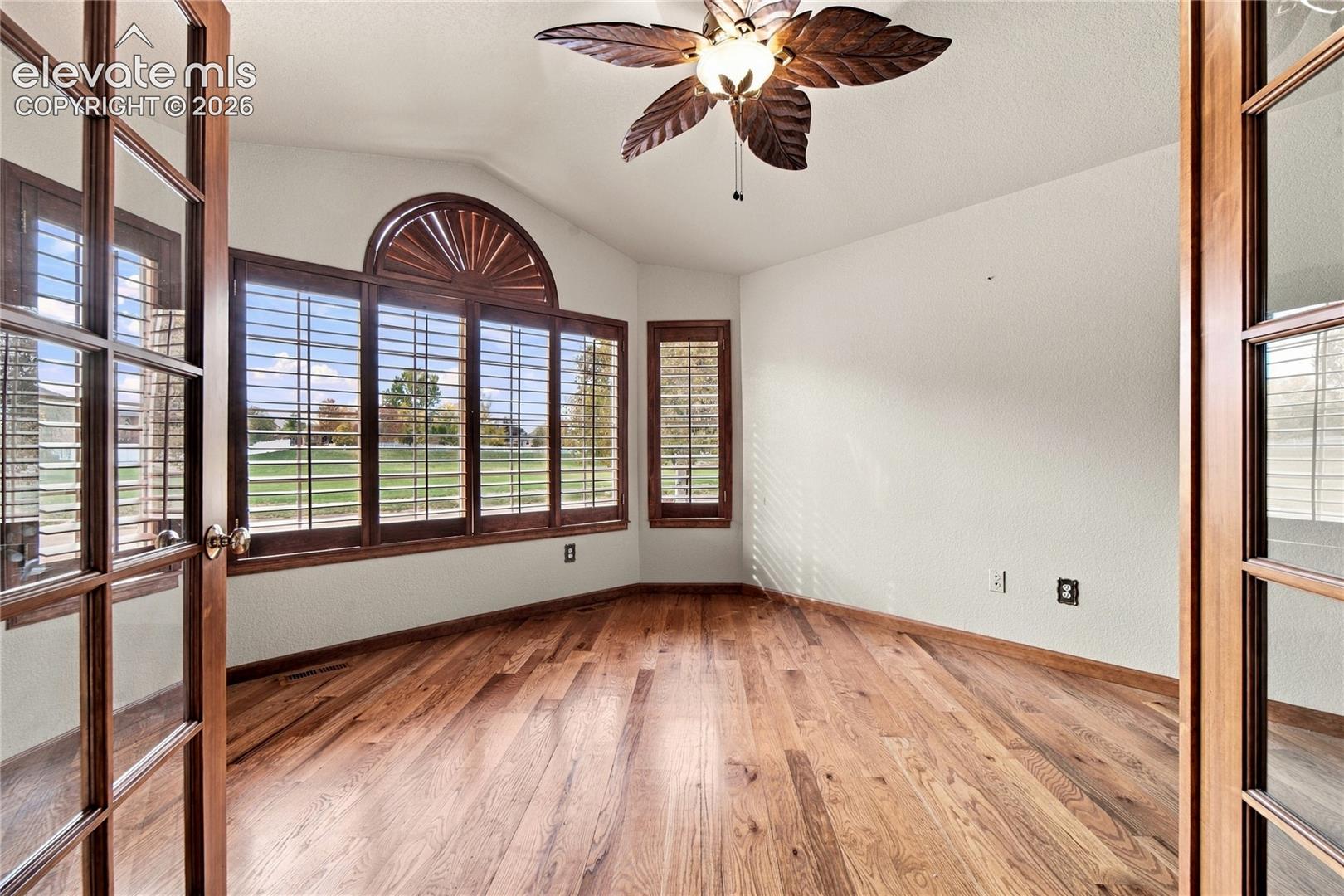 Wood-finish flooring, French doors with glass panes, large bay windows with arch detail, plantation shutters, and a ceiling fan with leaf-shaped blades