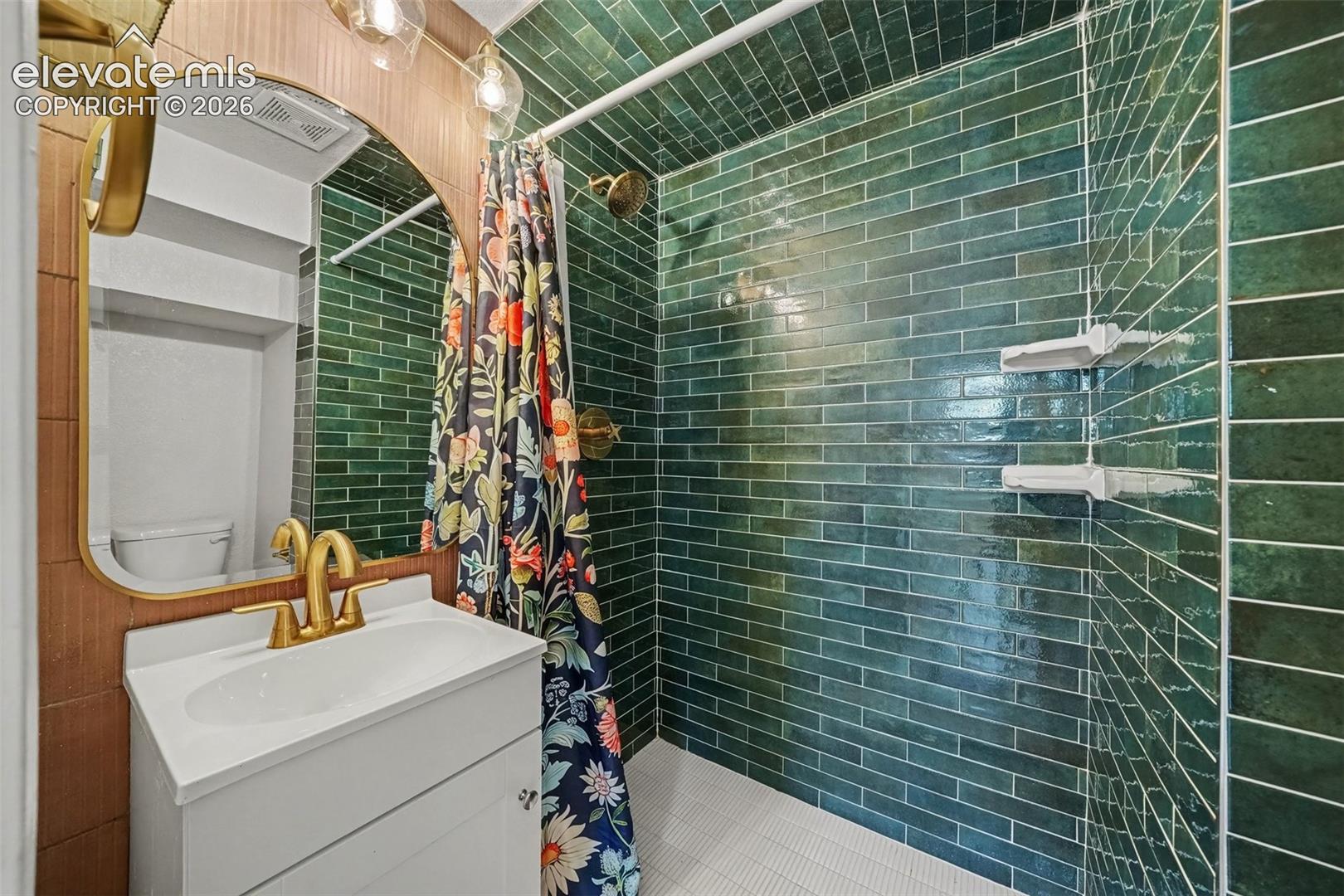 Bathroom featuring an arched vanity mirror with a brass frame, white cabinet vanity, brass finish faucet, and shower stall with green subway-style tile and built-in shelving