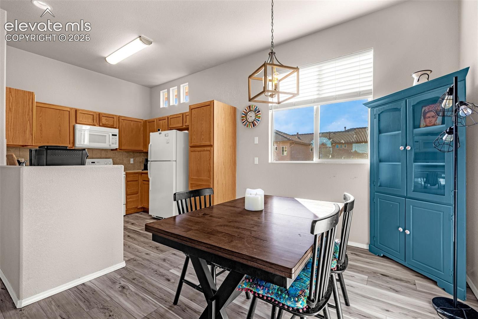 Kitchen featuring wood cabinetry, white appliances, wood-finish flooring, a geometric pendant light fixture, and large windows with horizontal blinds