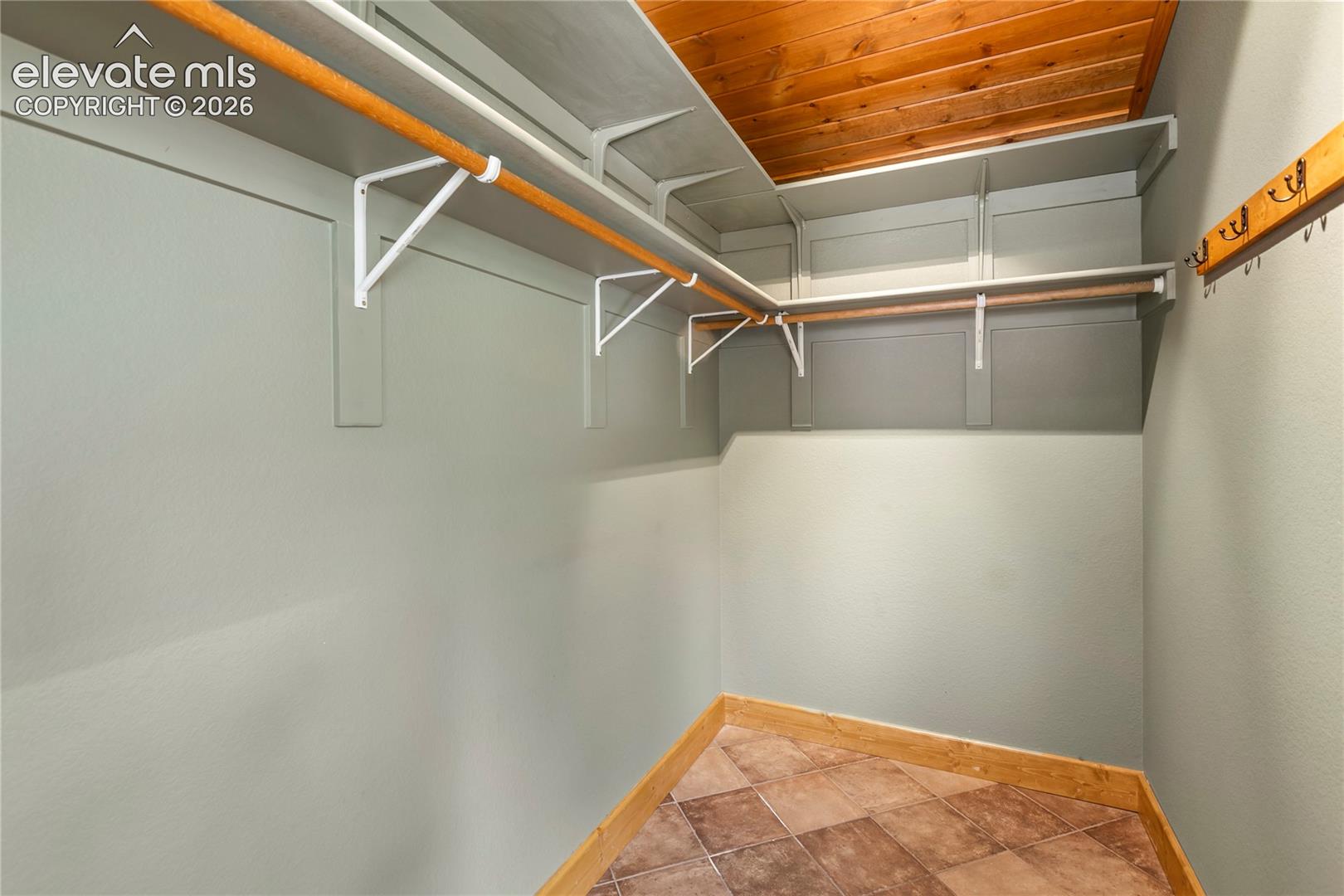 Storage space featuring tiered shelving with integrated garment rods, wood-paneled ceiling, ceramic tile flooring, and wall-mounted wooden hooks