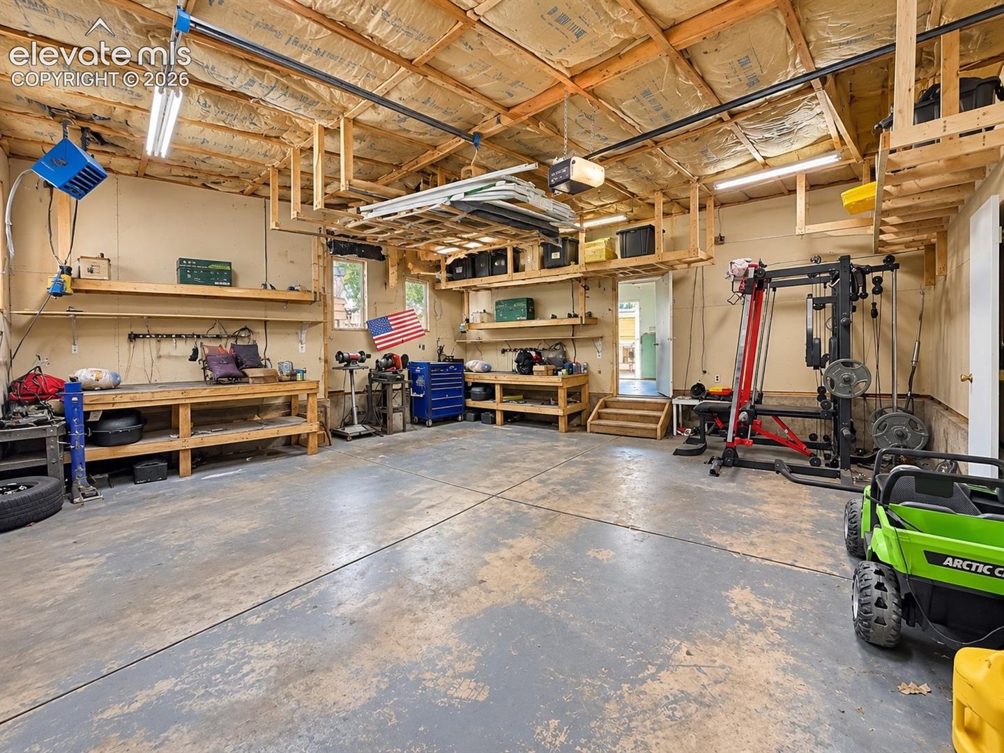 Spacious garage featuring a concrete floor, exposed wood beam ceiling, built-in shelving, elevated storage platforms, and a detached structure entrance with wood stairs